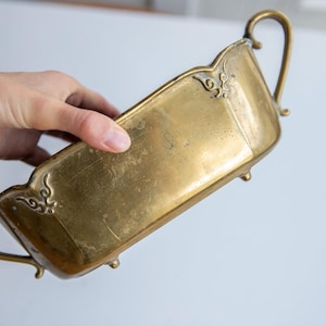 Pode incluir: Uma taça retangular de latão com acabamento polido, apresentando duas alças curvas nas laterais, quatro pés pequenos na base e detalhes decorativos em volutas em cada um dos quatro cantos superiores da borda do recipiente.