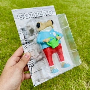 Könnte beinhalten: Figur einer Kreatur in einer transparenten Kunststoffverpackung. Die Figur trägt ein blaues Hemd, eine rote Hose und blaue Schuhe und hält eine grüne Gießkanne. Auf der Verpackung steht das Wort "Concho".