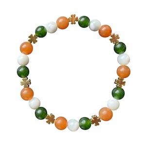 May include: A beaded bracelet featuring orange, white, and green beads, interspersed with gold-colored clover-shaped charms. The bracelet is strung on an elastic band, creating a circular shape. The beads are smooth and round, with a polished finish.