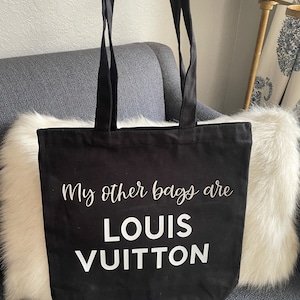 May include: A black canvas tote bag with two shoulder straps. The front of the bag features white text that reads "My other bags are LOUIS VUITTON" in a combination of cursive and block lettering.