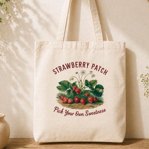 以下が含まれることがあります： クリーム色のトートバッグ。バーガンディ色で「Strawberry Patch」と「Pick Your Own Sweetness」の文字がプリントされています。イチゴと緑の葉のカラフルなイラストが特徴です。バッグには2つのハンドルが付いています。