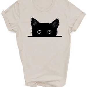 May include: A beige t-shirt with a black silhouette of a cat peeking over the top. The cat has large, white eyes.