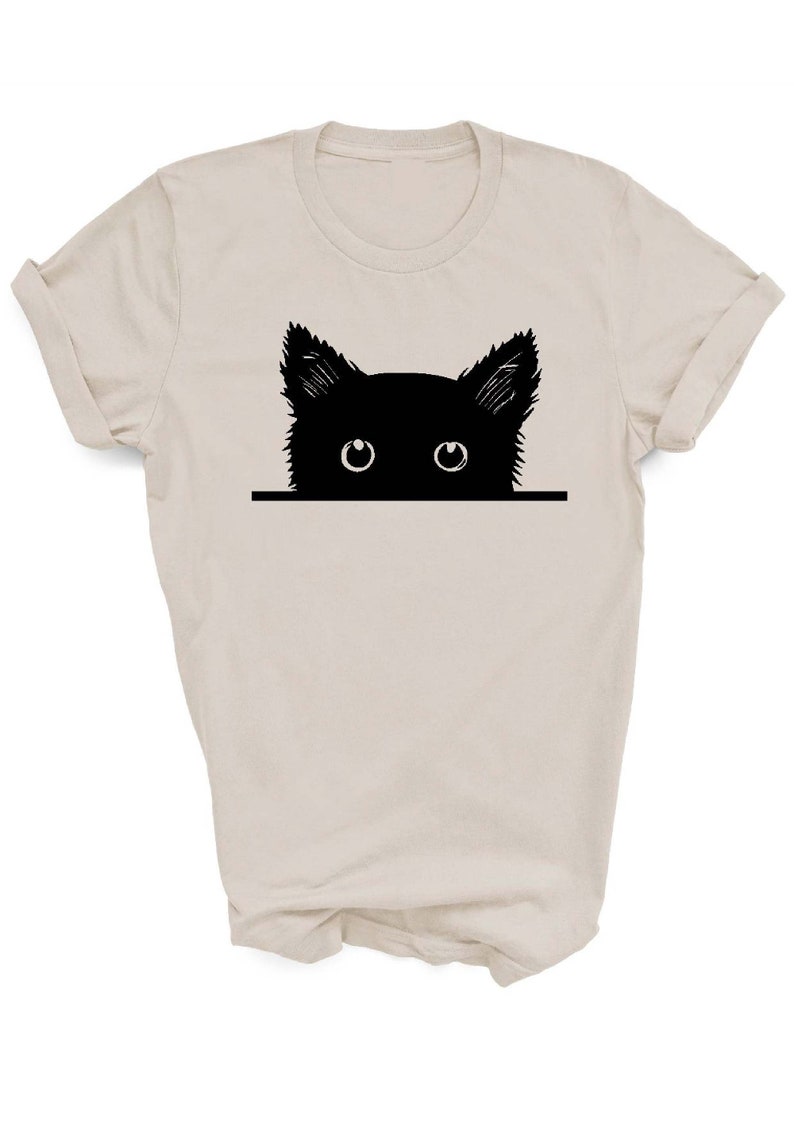 May include: A beige t-shirt with a black silhouette of a cat peeking over the top. The cat has large, white eyes.
