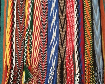 May include: A collection of colorful woven belts with intricate patterns. The belts are made with different colored threads and have a variety of geometric designs. The belts are all tied together at the bottom.