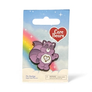 May include: A pin badge featuring a purple raccoon character from the Care Bears series. The pin is on a blue card with a rainbow and the Care Bears logo. The raccoon has a heart-shaped belly badge.