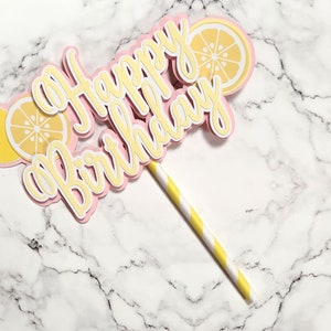 May include: A pink and yellow cake topper with the words "a Happy Birthday" and lemon slices. The topper is attached to a yellow and white striped straw.