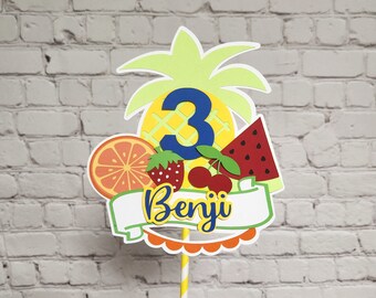 May include: A yellow and green pineapple cake topper with a blue number 3 and the name Benji on a white banner. The topper is surrounded by fruit cutouts, including a red strawberry, a red cherry, an orange slice, and a watermelon slice.