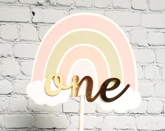May include: A pink, beige, and gold rainbow cake topper with the word "one" in gold script. The topper is on a white stick and is held in a hand.