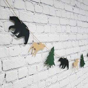 May include: A black, brown and green paper garland with cutouts of bears, moose and pine trees. The garland is strung on twine and is hanging on a white brick wall.