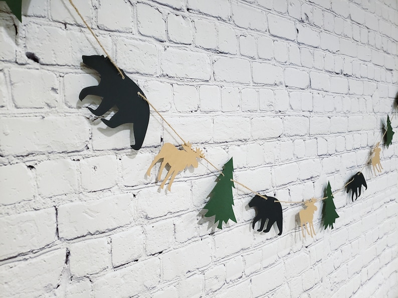 May include: A black, brown and green paper garland with cutouts of bears, moose and pine trees. The garland is strung on twine and is hanging on a white brick wall.