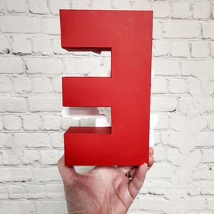 以下が含まれることがあります： 白いレンガの壁の前に、大きな赤い3次元の文字「E」が掲げられています。 この文字は、大胆でブロック状のデザインで、切り抜かれたセクションがあります。 白い雲の形をしたアクセントが文字の開口部に見えます。
