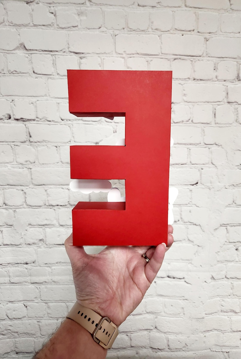 以下が含まれることがあります： 白いレンガの壁の前に、大きな赤い3次元の文字「E」が掲げられています。 この文字は、大胆でブロック状のデザインで、切り抜かれたセクションがあります。 白い雲の形をしたアクセントが文字の開口部に見えます。