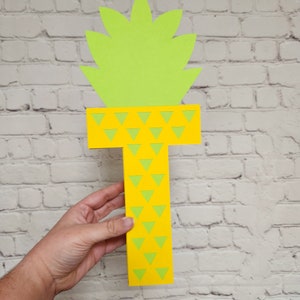 May include: A yellow paper letter T with a green pineapple top. The letter is decorated with green triangles.