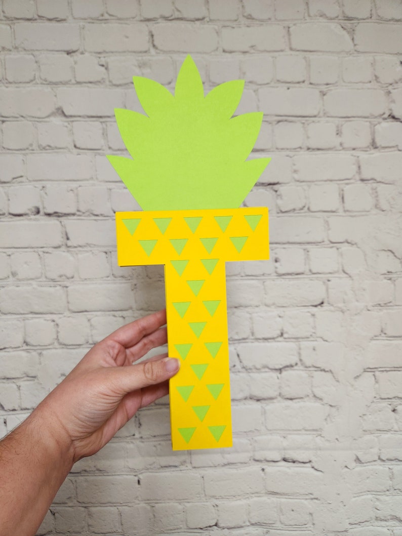 May include: A yellow paper letter T with a green pineapple top. The letter is decorated with green triangles.