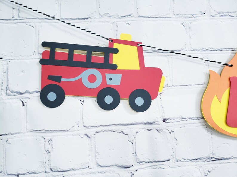 May include: Red fire truck paper cut-out with black wheels, a gray hose, and a yellow ladder. The fire truck is part of a paper banner with a yellow and orange flame cut-out.