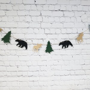 May include: A black, green and tan paper garland with alternating bear, moose and pine tree shapes. The garland is strung on a white brick wall background.