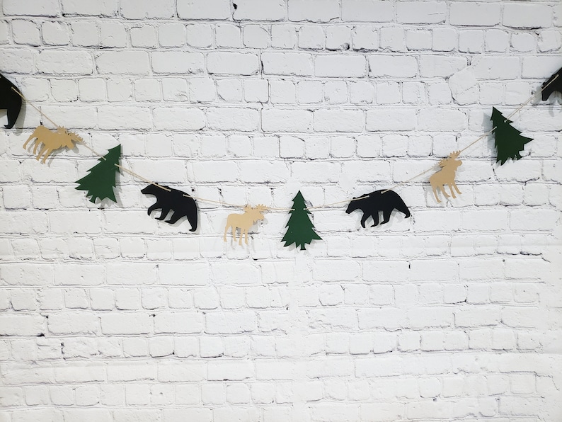 May include: A black, green and tan paper garland with alternating bear, moose and pine tree shapes. The garland is strung on a white brick wall background.