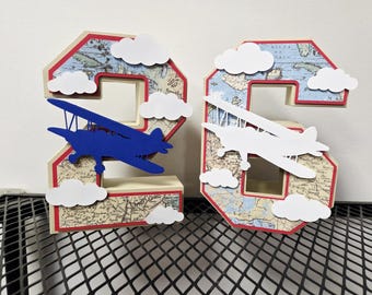 May include: Two decorative numbers, a 2 and a 6, with an aviation theme. Each number is made of a map design with a red border, white clouds, and a biplane. The number 2 has a blue biplane, and the number 6 has a white biplane.