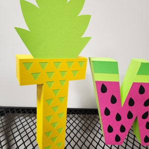 May include: Two large cardboard letters, "T" and "W", decorated with a pineapple and watermelon theme. The "T" is yellow with green triangle patterns and a green pineapple top. The "W" is pink with black seeds and a green watermelon rind.