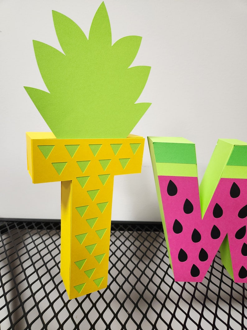 May include: Two large cardboard letters, "T" and "W", decorated with a pineapple and watermelon theme. The "T" is yellow with green triangle patterns and a green pineapple top. The "W" is pink with black seeds and a green watermelon rind.