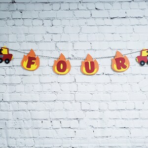 May include: Fire truck themed banner with red letters spelling out "FOUR" in a flame design. The banner is strung with red and white string and has a fire truck on each end.