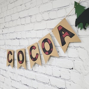 May include: A brown paper banner with the words "HOT COCOA" in black and red buffalo check letters. The banner is strung with twine and has a green paper tree and a black paper bear hanging from it.