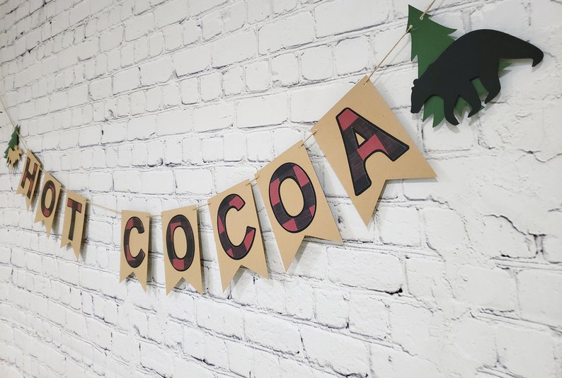 May include: A brown paper banner with the words "HOT COCOA" in black and red buffalo check letters. The banner is strung with twine and has a green paper tree and a black paper bear hanging from it.