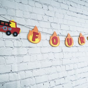 May include: Fire truck and flame-shaped letters spelling out "FOUR" on a white brick wall. This is a birthday banner for a child's fourth birthday party.