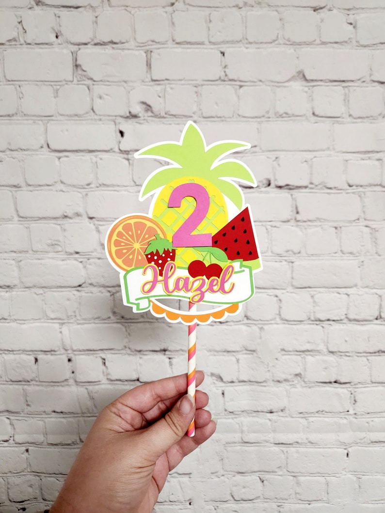 K&ouml;nnte beinhalten: Ein rosa Nummer-Zwei-Kuchentopper mit Ananas, Wassermelone, Orange, Erdbeere und Kirschen. Der Topper hat ein Banner mit der Aufschrift "Hazel" in rosa Buchstaben.