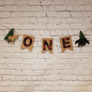 May include: A brown and red "ONE" banner with a moose and a bear silhouette, decorated with green pine trees. The banner is hanging on a white brick wall.