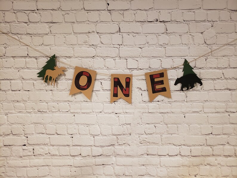 May include: A brown and red "ONE" banner with a moose and a bear silhouette, decorated with green pine trees. The banner is hanging on a white brick wall.