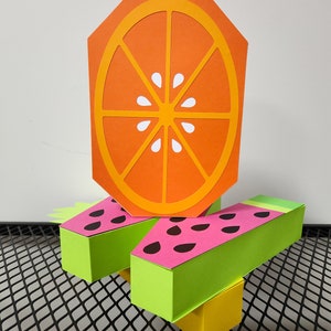 May include: A three-dimensional paper craft of a watermelon and orange. The watermelon is green and pink with black seeds. The orange is orange with a white center.