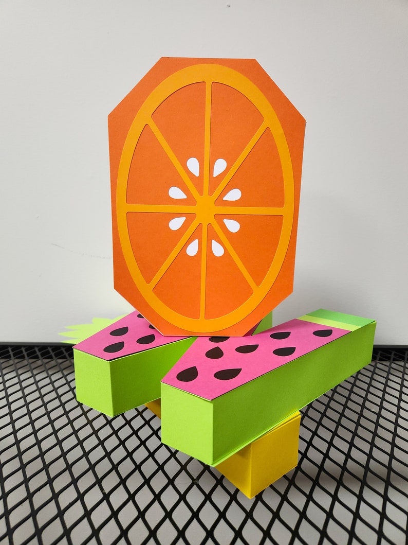 May include: A three-dimensional paper craft of a watermelon and orange. The watermelon is green and pink with black seeds. The orange is orange with a white center.