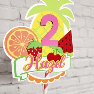 K&ouml;nnte beinhalten: Ein bunter Tortendekor mit Ananas, Wassermelone, Orange, Erdbeere und Kirschen. Die Zahl 2 ist in Pink und der Name Hazel ist in Pink und Gelb. Der Topper ist auf einem gestreiften Strohhalm.
