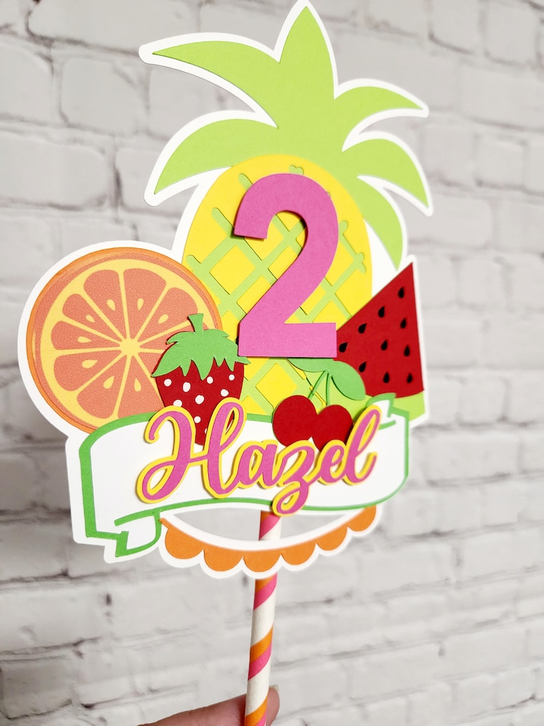 K&ouml;nnte beinhalten: Ein bunter Tortendekor mit Ananas, Wassermelone, Orange, Erdbeere und Kirschen. Die Zahl 2 ist in Pink und der Name Hazel ist in Pink und Gelb. Der Topper ist auf einem gestreiften Strohhalm.