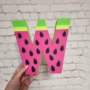 May include: A large, pink and green cardboard letter W shaped like a watermelon. The letter has black seed details.