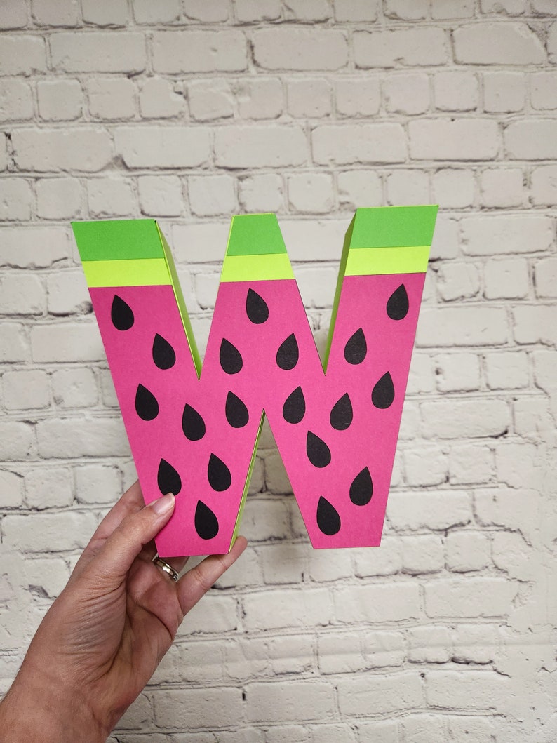 May include: A large, pink and green cardboard letter W shaped like a watermelon. The letter has black seed details.