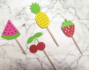 May include: Four paper fruit cupcake toppers on wooden sticks. The toppers are a watermelon, a pineapple, a cherry, and a strawberry. The watermelon is pink with black seeds, the pineapple is yellow with green leaves, the cherry is red with green leaves, and the strawberry is red with white seeds.