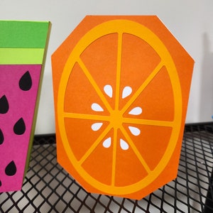 May include: Orange slice cutout made of orange and yellow paper with a white center. The cutout is on a black wire mesh surface.