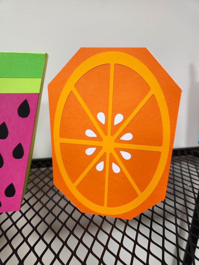 May include: Orange slice cutout made of orange and yellow paper with a white center. The cutout is on a black wire mesh surface.