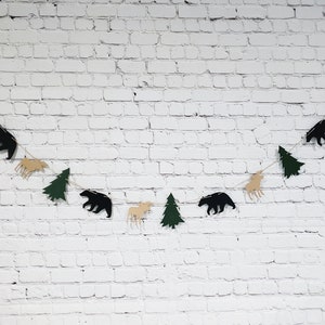 May include: A green and brown paper garland with cutouts of trees, moose, and bears. The garland is strung on a white brick wall.