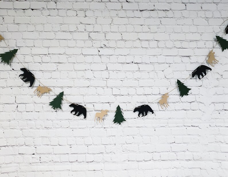 May include: A green and brown paper garland with cutouts of trees, moose, and bears. The garland is strung on a white brick wall.