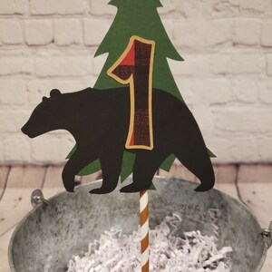 May include: A black bear silhouette cake topper with a green pine tree and a red and black checked number one. The topper is on a brown and white striped stick.
