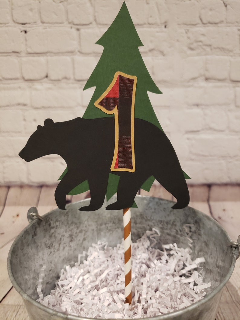 May include: A black bear silhouette cake topper with a green pine tree and a red and black checked number one. The topper is on a brown and white striped stick.