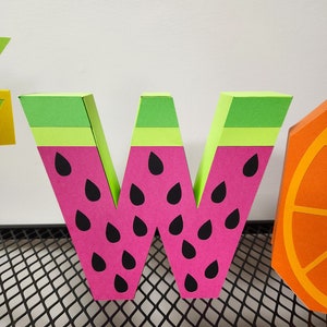 May include: A pink and green paper letter W with black watermelon seeds. The letter is standing on a black wire mesh surface.