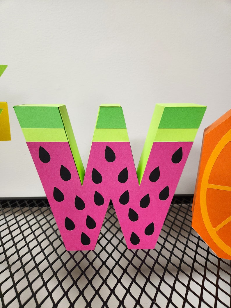 May include: A pink and green paper letter W with black watermelon seeds. The letter is standing on a black wire mesh surface.