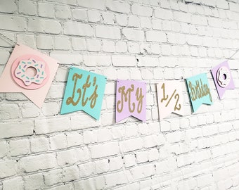 May include: A pastel-colored banner with the words "It's My 1/2 Birthday" in gold glitter lettering. The banner is decorated with two pink and white donut cutouts.
