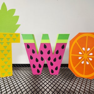 May include: The word "TWO" is spelled out in colorful paper letters. The letter "T" is yellow and has a pineapple design. The letter "W" is pink and has a watermelon design. The letter "O" is orange and has an orange slice design.