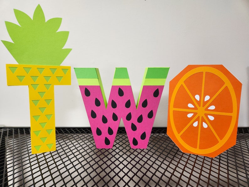 May include: The word "TWO" is spelled out in colorful paper letters. The letter "T" is yellow and has a pineapple design. The letter "W" is pink and has a watermelon design. The letter "O" is orange and has an orange slice design.