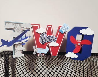 May include: A decorative 'TWO' sign with an airplane theme. The letters are blue, red, and white, with cloud and airplane cutouts. The letter 'W' has the name 'Owen' on it. Perfect for a birthday.